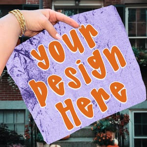 May include: A light purple textile with the words "YOUR DESIGN HERE" in orange letters. The textile has a distressed, textured appearance. A hand with a gold bracelet points to the text.