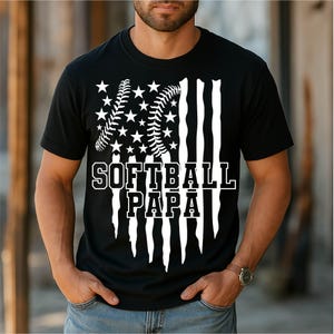 Op de afbeelding: Zwart t-shirt met een wit grafisch ontwerp. Het ontwerp toont een softbal en sterren die zijn gerangschikt om op de Amerikaanse vlag te lijken, met de woorden "SOFTBALL PAPA" in vette letters.