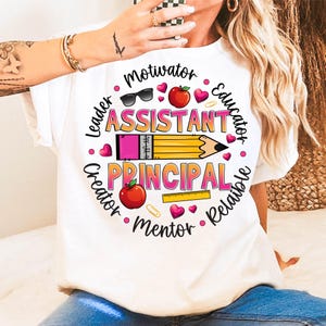 May include: White t-shirt with a colorful graphic design for an assistant principal. The design includes a pencil, apple, sunglasses, and the words "Motivator, Leader, Creator, Mentor, Reliable, Educator." The word "Assistant Principal" is prominently displayed.