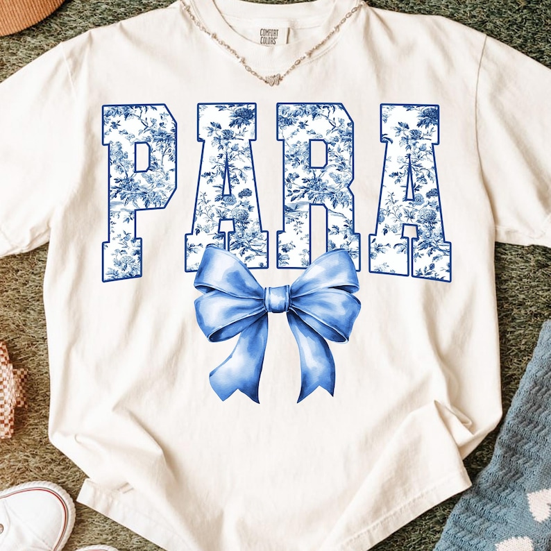 May include: Cream-colored t-shirt with the word "PARA" in blue floral print letters and a blue bow graphic. The shirt has a crew neck and short sleeves. A silver necklace is visible at the neckline.