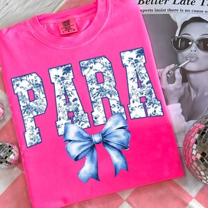 May include: A bright pink t-shirt with the word "PARA" in large, white letters filled with a blue floral pattern. A blue bow is centered below the text. The shirt is on a pink and white checkered surface.