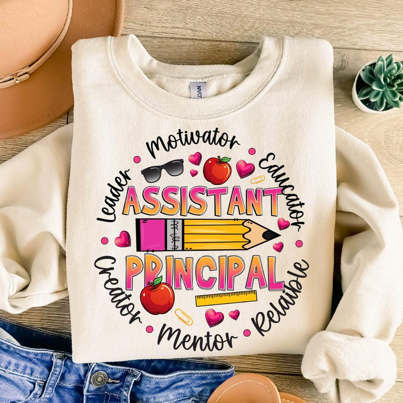 May include: Cream-colored sweatshirt with a colorful graphic design. The design features the words "Assistant Principal" in pink and orange, surrounded by words like "Leader" and "Educator." A large pencil graphic is in the center, with hearts, apples, and sunglasses.