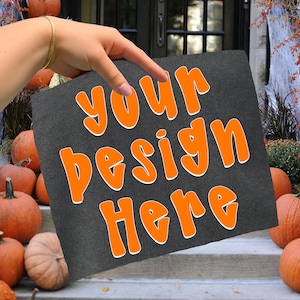 Pode incluir: Um quadrado cinza escuro com as palavras "your design here" em laranja, contornadas em branco. O quadrado é segurado em frente a um fundo de abóboras e uma porta.