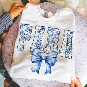 May include: White sweatshirt with the word "PARA" in blue floral print letters and a blue bow graphic. The sweatshirt is held by hands, and the background is a floral pattern.