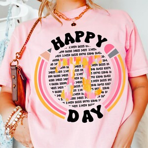 May include: Pink t-shirt with a rainbow design and the text "HAPPY DAY" in a retro font. The rainbow is made up of colorful stripes with numbers and mathematical symbols inside each stripe.