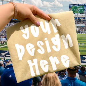 Puede incluir: Camiseta color beige con el texto blanco "your design here", sostenida frente a una multitud en un estadio. La camiseta es de color neutro y el texto es en negrita y fácil de leer. El fondo muestra un partido de fútbol.