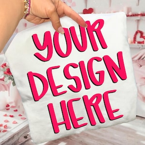 May include: White pillow with the text "YOUR DESIGN HERE" in bright pink, outlined in black. The pillow is held up by a hand with a bracelet. The background includes Valentine's Day decorations.