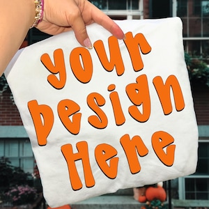 May include: White pillow with the text "your design here" in orange bubble letters. The pillow is being held up by a hand with a gold bracelet. The background is a brick building with a window.