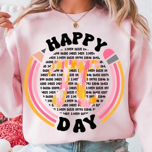 May include: A pink sweatshirt with a rainbow design and the text "HAPPY DAY" in a retro font. The rainbow is made up of numbers and the text is surrounded by a yellow circle.