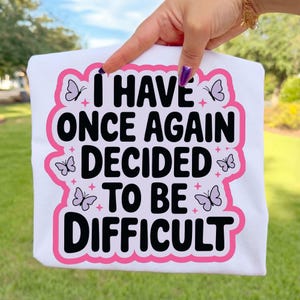 May include: White t-shirt with the phrase "I HAVE ONCE AGAIN DECIDED TO BE DIFFICULT" in black text, outlined in pink. The design includes butterfly illustrations and star accents.