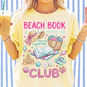 May include: Yellow t-shirt featuring a beach-themed design with a sun hat, books, and seashells. The text reads "Beach Book Club". The shirt has a relaxed fit and short sleeves.