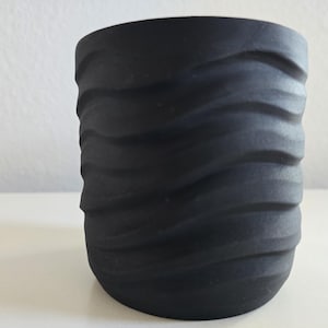 May include: A matte black cylindrical planter featuring a series of horizontal, undulating wave-like ridges carved into its exterior surface. The vessel has a wide, open top and a rounded base, presenting a uniform dark finish throughout the design.
