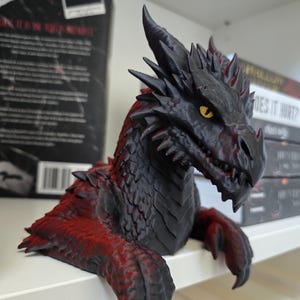 May include: A dragon figurine featuring a black and red color scheme with sharp, textured scales, prominent horns, and piercing yellow eyes. The creature is depicted in a forward-leaning pose with its front claws resting over the edge of a surface.