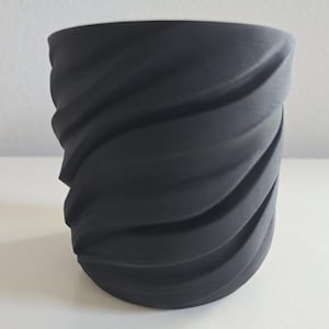 May include: A matte black cylindrical planter featuring a distinct spiraling, wave-like texture across its exterior surface. The vessel has a wide, open top and a tapered base, showcasing a uniform dark finish with deep, recessed grooves.