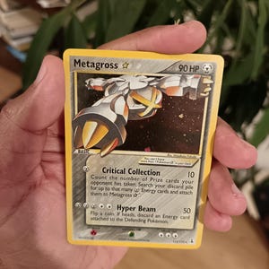 Puede incluir: Una carta coleccionable Metagross Star con 90 HP, que muestra una criatura metálica plateada y dorada sobre un fondo oscuro estrellado. El texto de la carta incluye los ataques Critical Collection y Hyper Beam, con el número 113/113 en la parte inferior.
