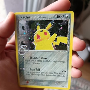 Puede incluir: Una carta coleccionable Pikachu Delta Species con 40 HP. La carta presenta una ilustración amarilla de Pikachu sobre un fondo oscuro holográfico. El texto incluye los ataques Thunder Wave e Iron Tail, con información básica y el crédito de Ryo Ueda.