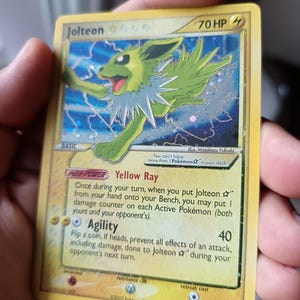 Puede incluir: Una carta coleccionable de Jolteon con 70 HP, que muestra una ilustración de personaje amarillo y verde sobre un fondo de rayos azules. La carta incluye el texto Yellow Ray y Agility, con un acabado holográfico en un marco amarillo.