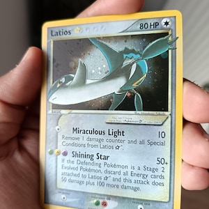 Puede incluir: Una carta coleccionable Pokémon Latios Star con 80 HP, que presenta una ilustración de una criatura azul y blanca sobre un fondo oscuro estrellado. El texto de la carta incluye los ataques Miraculous Light y Shining Star, con fecha de 2005.