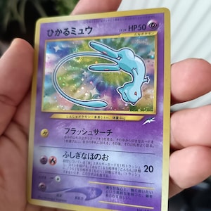 Puede incluir: Una carta coleccionable Pokémon Mew brillante japonesa con una ilustración holográfica de la criatura azul claro sobre un fondo morado. La carta muestra 50 HP, nivel 24 y texto en japonés, incluido el nombre Hikaru Mew.