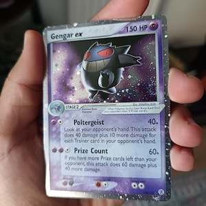 Puede incluir: Una carta coleccionable Gengar ex con 150 HP, que presenta un fondo holográfico morado y al personaje Gengar. La carta muestra los ataques Poltergeist y Prize Count, con texto que detalla los mecanismos de daño y la evolución desde Haunter.