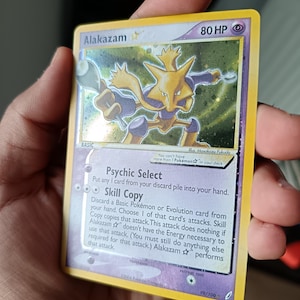 Puede incluir: Una carta coleccionable Pokémon Alakazam con borde amarillo y 80 HP, que presenta una ilustración holográfica del personaje psíquico sosteniendo dos cucharas plateadas. El texto incluye las habilidades Psychic Select y Skill Copy, numerada 99/100.