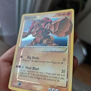 Puede incluir: Una carta coleccionable Regirock Star con 90 HP, que muestra una ilustración de una criatura rocosa marrón. El texto de la carta incluye los ataques Dig Drain y Final Blast, fecha de copyright 2006, borde amarillo y acabado holográfico.