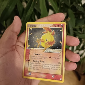 Puede incluir: Una carta coleccionable Pokémon Torchic Gold Star con 70 HP. La carta muestra un personaje de pollito amarillo sobre un fondo oscuro y estrellado. El texto incluye los ataques Fireworks y Spring Back, el número 108/109 y el año de copyright 2004.