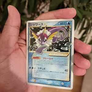 Pode incluir: Uma carta colecionável japonesa Vaporeon Gold Star apresentando o personagem Pokémon roxo com uma barbatana amarela, cercado por bolhas de água. A carta exibe o nome シャワーズ, HP70 e texto em japonês detalhando suas habilidades.