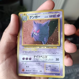 Op de afbeelding: Een Japanse Gengar Pokemon-verzamelkaart met een paars spookachtig personage met een brede grijns. De kaart toont HP80, level 38, en bevat Japanse tekst, met een holografisch achtergrondpatroon achter de centrale illustratie.