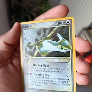 Puede incluir: Una carta coleccionable Pokémon Latias Star con 80 HP, que presenta una ilustración de una criatura blanca y verde. El texto de la carta incluye los ataques Healing Light y Shooting Star, con fecha de copyright 2005 y número 105/107 abajo.