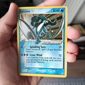 Puede incluir: Una carta coleccionable Pokémon Suicune Star con 80 HP, con un diseño en tonos azules y blancos. La carta muestra los ataques Splashing Turn y Cross Wind, junto con texto sobre las reglas del juego, copyright de 2005 e ilustración holográfica.