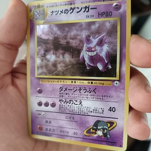 Peut inclure: Une carte à collectionner japonaise violette représentant le personnage Gengar, étiquetée Natsume no Gengar, avec 80 PV, niveau 39, et une illustration holographique de la créature de type spectre dans un environnement sombre et brumeux.