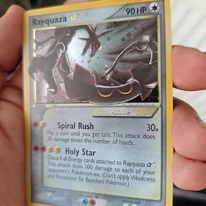Puede incluir: Una carta coleccionable Rayquaza Gold Star con 90 HP, que presenta una ilustración de una criatura negra similar a un dragón. El texto incluye los ataques Spiral Rush y Holy Star, el crédito del ilustrador Masakazu Fukuda y el número 107/107.
