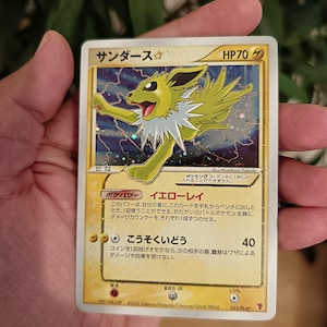 Può includere: Una carta collezionabile Pokémon giapponese Jolteon Gold Star raffigurante una creatura gialla con un colletto bianco a punta su uno sfondo blu scuro stellato. La carta mostra HP70, simboli di energia elettrica e testo in giapponese.