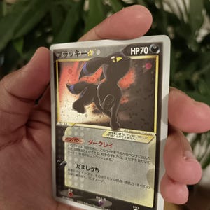 Può includere: Una carta collezionabile Pokémon giapponese Umbreon Gold Star raffigurante una creatura nera con occhi gialli e dettagli blu su sfondo scuro. La carta mostra 70 HP, il nome "ブラッキー" e testo in giapponese che descrive abilità e statistiche.