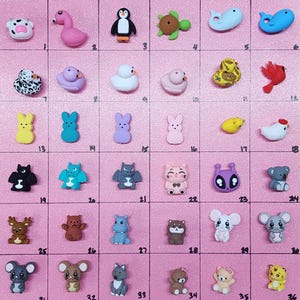 May include: A set of 36 colorful resin flatback cabochons featuring various animal, food, and holiday themes. The cabochons are arranged in a grid pattern on a pink background.