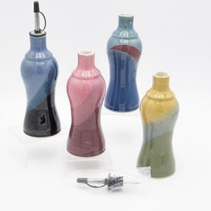 Op de afbeelding: Vier keramische olieflessen in blauw, roze en geel. Eén fles heeft een roestvrijstalen tuit. De flessen hebben een gebogen vorm en worden op transparante standaards getoond. Een klein pakketje tuiten is ook zichtbaar.