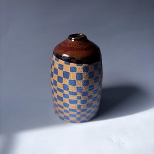May include: A small, handcrafted ceramic vase. It has a brown top and a body decorated with a blue and tan checkerboard pattern. The vase is a decorative item.
