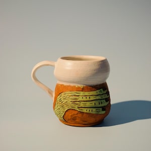 Pode incluir: Caneca de cerâmica artesanal com borda e alça creme. O corpo da caneca é laranja com um design texturizado verde. A caneca é projetada para bebidas quentes e é adequada para uso doméstico ou no escritório.