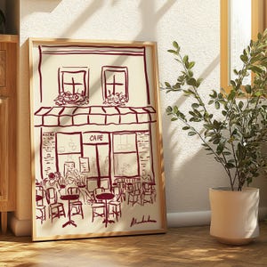 Könnte beinhalten: Ein gerahmter Druck einer Cafészene im minimalistischen Stil. Das Kunstwerk zeigt eine bordeauxfarbene Strichzeichnung eines Café-Äußeren mit Tischen, Stühlen und Personen. Das Wort "CAFE" ist über dem Eingang zu sehen.