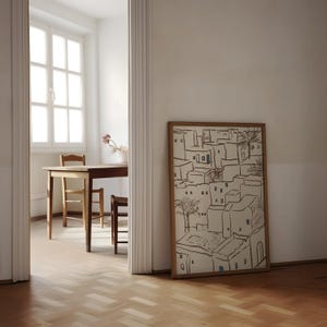 Pu&ograve; includere: Un'opera d'arte incorniciata raffigurante una scena di villaggio con edifici beige, finestre blu e palme. L'opera &egrave; in una cornice di legno naturale e si appoggia a una parete bianca. La stanza ha un pavimento in legno e uno scorcio di un tavolo e sedie.