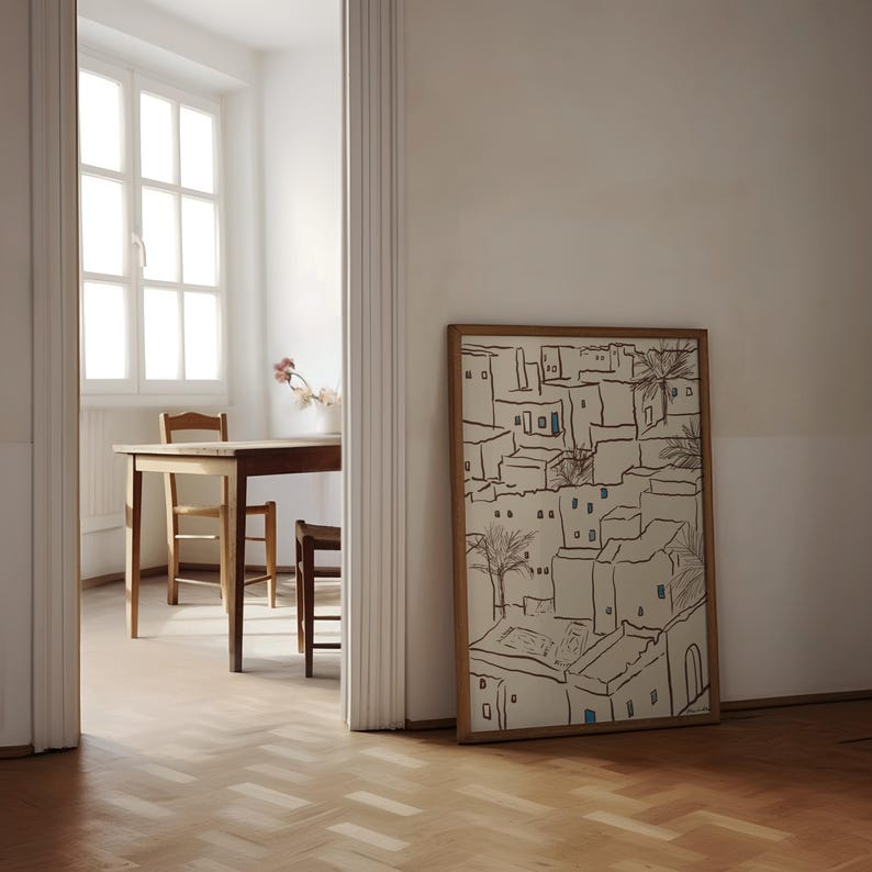 Pu&ograve; includere: Un'opera d'arte incorniciata raffigurante una scena di villaggio con edifici beige, finestre blu e palme. L'opera &egrave; in una cornice di legno naturale e si appoggia a una parete bianca. La stanza ha un pavimento in legno e uno scorcio di un tavolo e sedie.