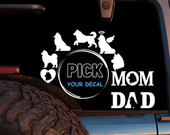 May include: A black car window decal featuring white silhouettes of various dog breeds. The text includes "PICK YOUR DECAL", "MOM", and "DAD". The design incorporates a heart and an angel dog.
