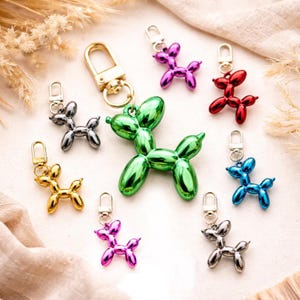 May include: A collection of metallic balloon dog keychains in various colors, including gold, silver, green, red, blue, and purple. Each keychain features a lobster clasp for easy attachment. The keychains are arranged on a light-colored surface.