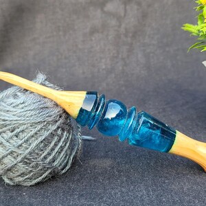 May include: A wooden crochet hook with a blue resin handle. The hook is lying on a ball of gray yarn.