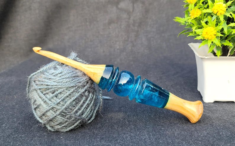 May include: A wooden crochet hook with a blue resin handle. The hook is lying on a ball of gray yarn.