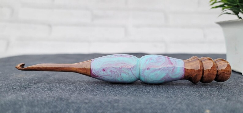 May include: A wooden crochet hook with a blue and pink marbled handle. The hook is made of wood and has a smooth finish.