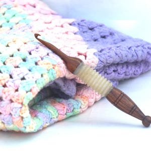 May include: A close-up of a wooden crochet hook with a light purple and white handle. The hook is lying on top of a multi-colored crochet blanket.