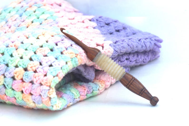 May include: A close-up of a wooden crochet hook with a light purple and white handle. The hook is lying on top of a multi-colored crochet blanket.