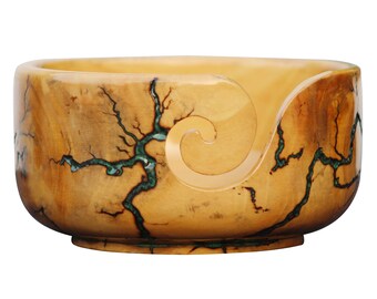 Könnte beinhalten: Eine Holzwolle Schüssel mit einem einzigartigen Wirbeldesign und einem natürlichen Holzmaserungsmuster. Die Schüssel hat eine hellbraune Farbe mit grünen Akzenten.